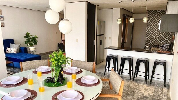 A dining area with a round table set for six, surrounded by chairs, and a kitchen area with a refrigerator and bar stools in the background. - HUGE. FULL APARTMENT 100% REMODELED EXPO AREA (Zapopan)