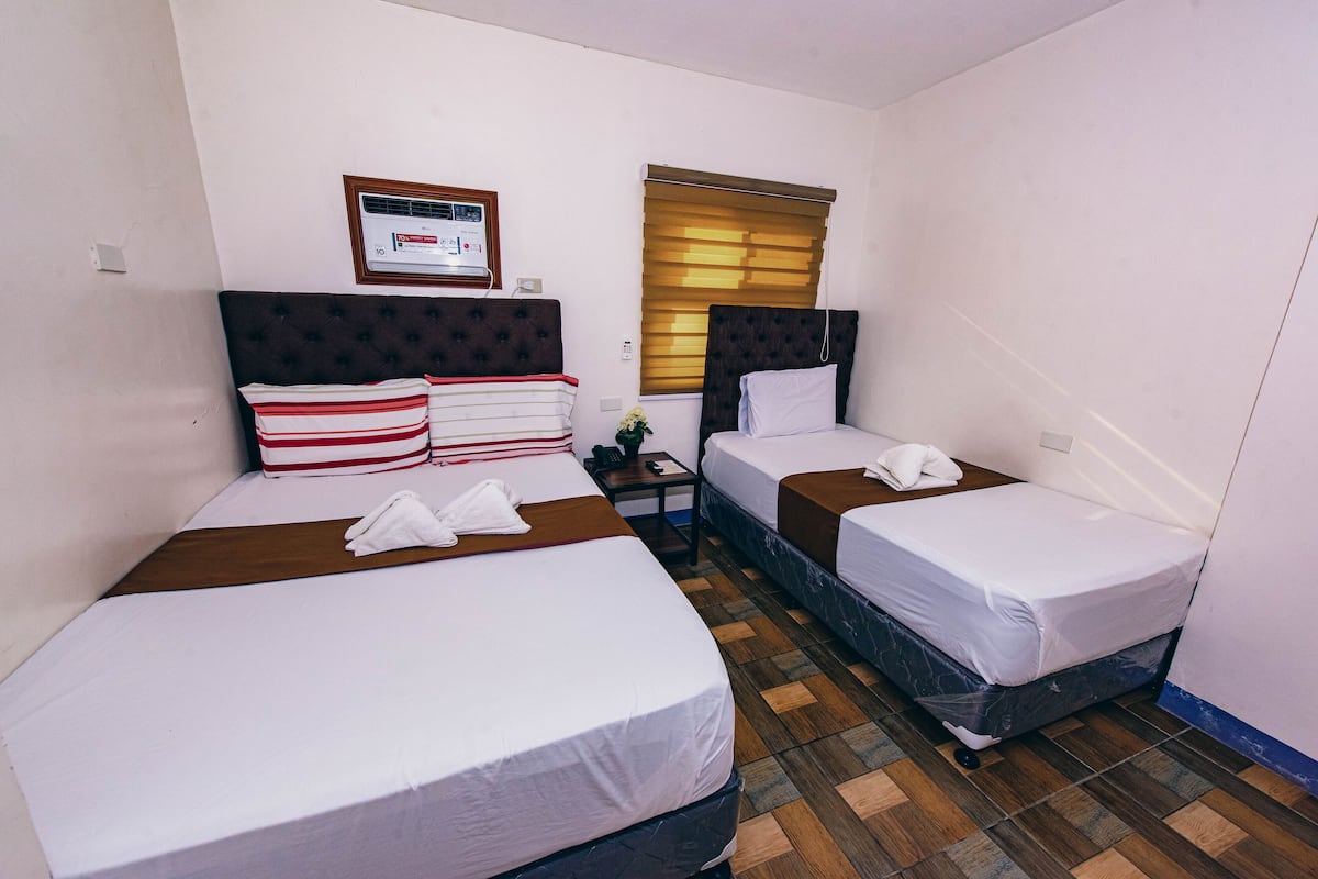 Quarto de hotel com duas camas, uma pequena mesa, uma mesinha de cabeceira, uma janela com persianas e um termostato na parede.