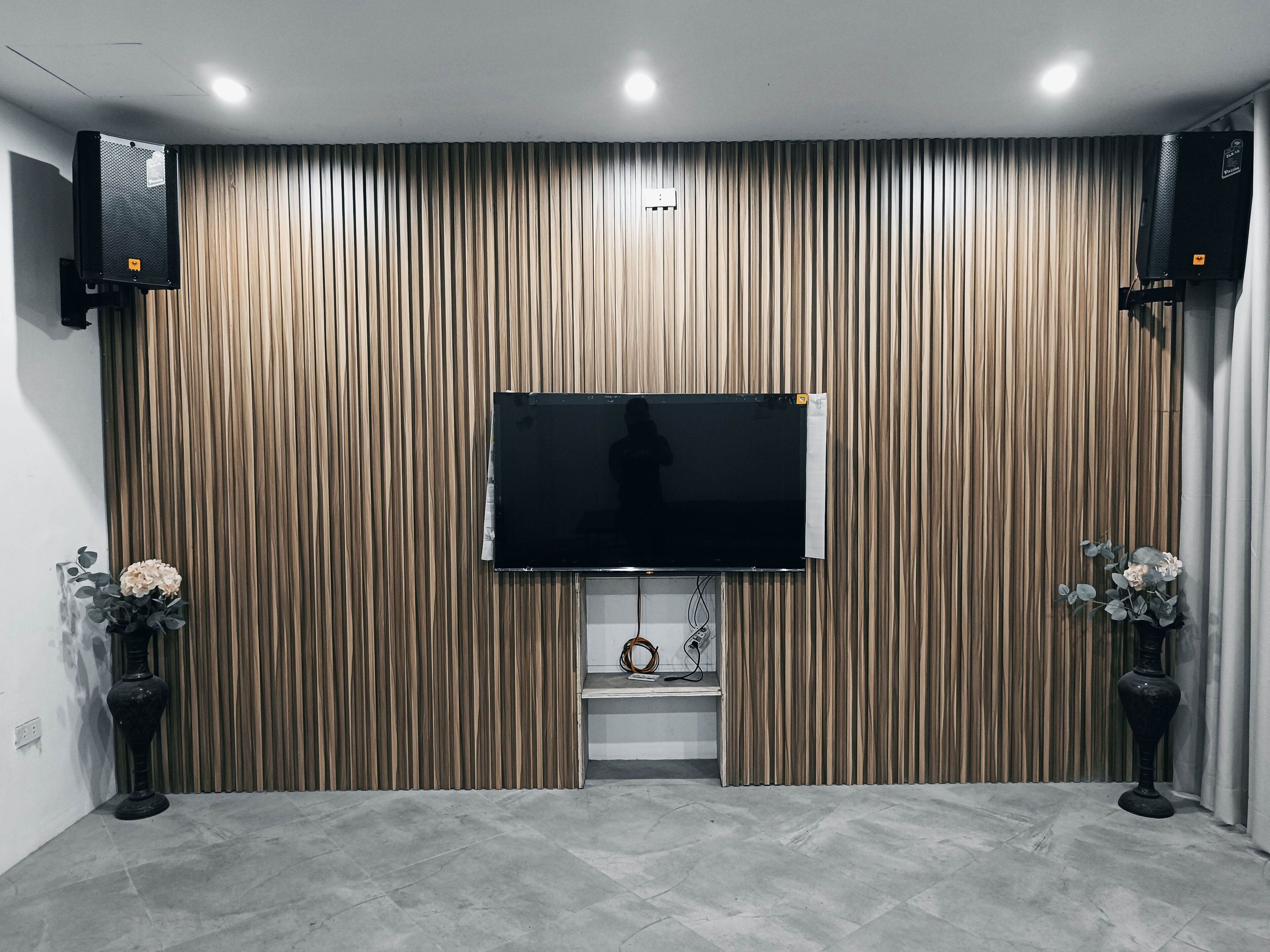A modern room with a flat-screen TV mounted on a wooden panel wall, two speakers on either side, and a small shelf with decorative items.