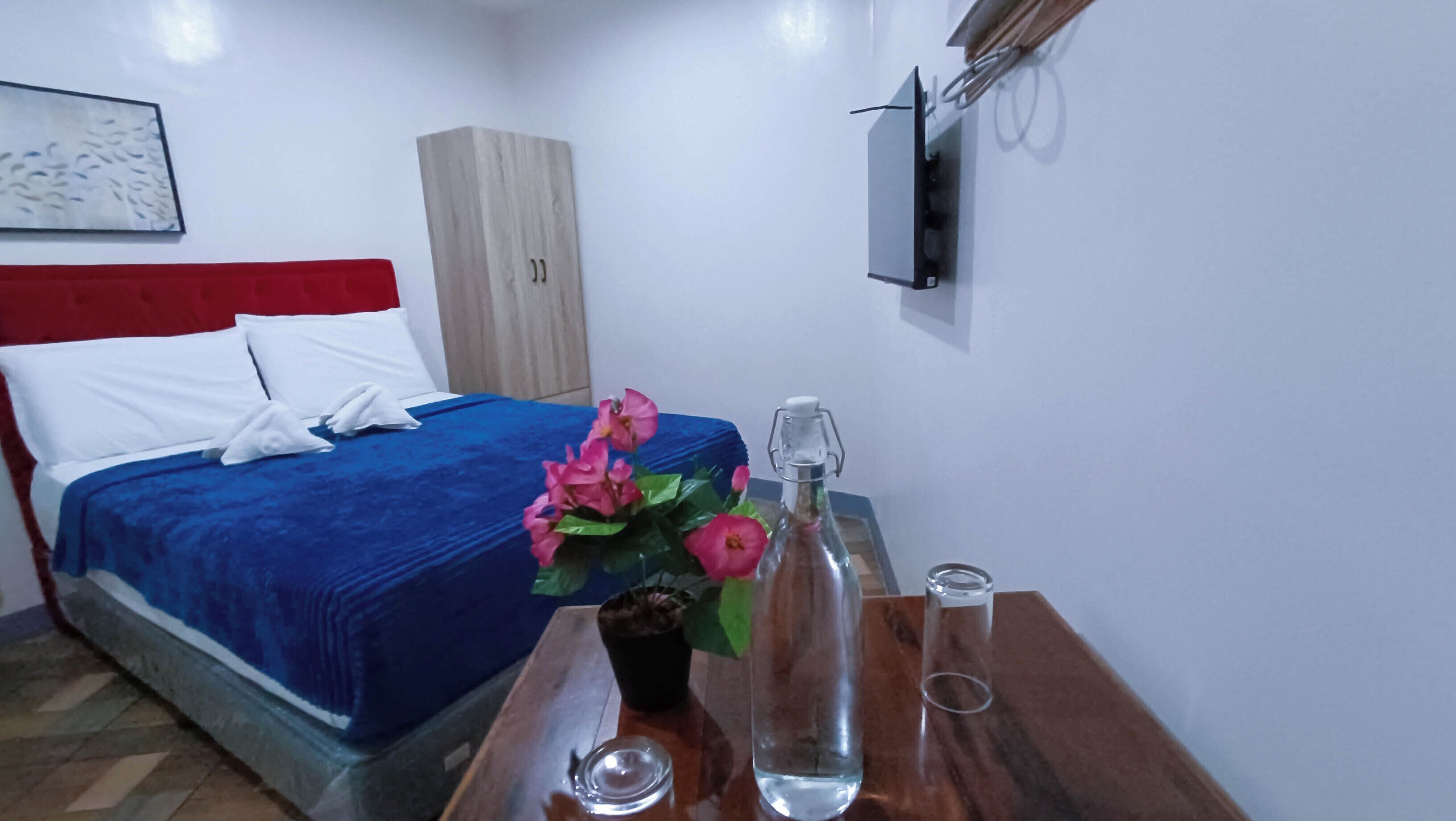 A hotel room with a bed, a wooden nightstand, a bottle of water, a glass, and a vase with flowers.