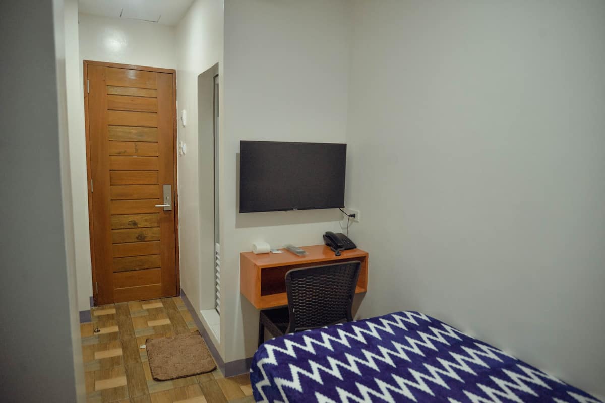 Um quarto pequeno com porta de madeira, uma escrivaninha com cadeira, uma cama com coberta estampada e uma TV presa na parede.