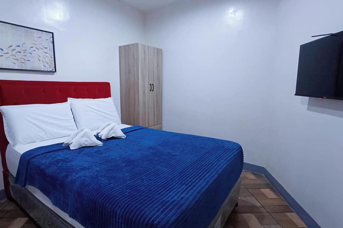 Cama bem arrumada com roupa de cama branca e azul, cabeceira vermelha e guarda-roupa de madeira.