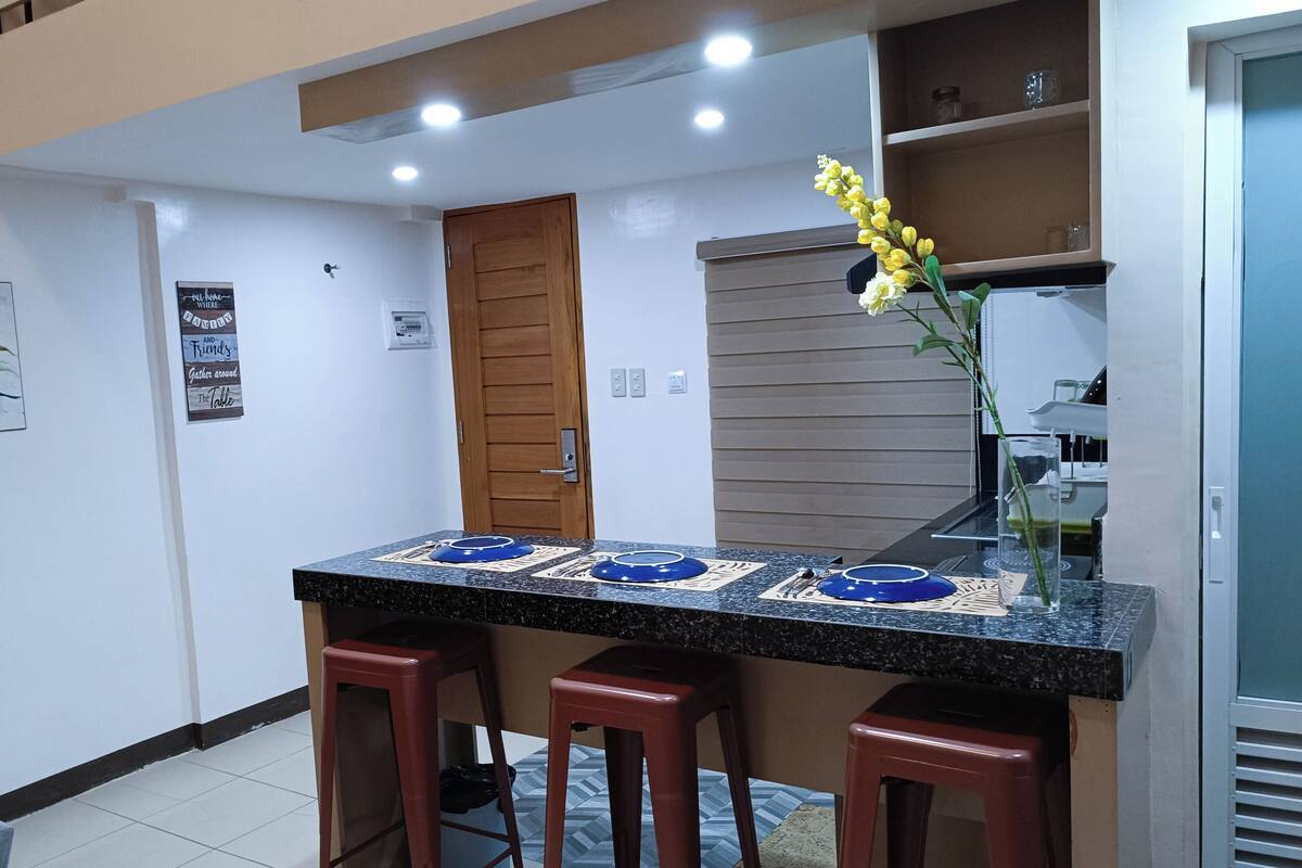 Cozinha moderna com bancada de granito, três banquetas, uma porta de madeira e uma pequena estante com objetos decorativos.