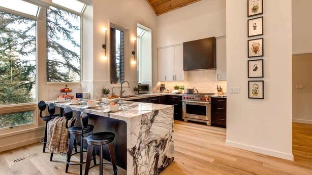 A modern kitchen with a marble island, bar stools, and a large window overlooking trees.