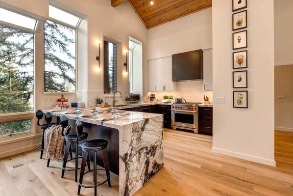A modern kitchen with a marble island, bar stools, and a large window overlooking trees.