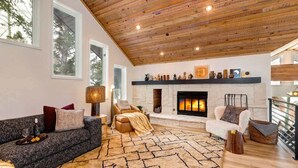 A spacious living room with a fireplace, a high wooden ceiling, and a large window.