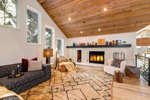 A spacious living room with a fireplace, a high wooden ceiling, and a large window.