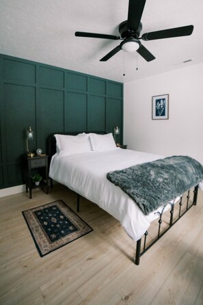 Un dormitorio con cama, ventilador de techo, mesita de noche, lámpara, un cuadro enmarcado en la pared y una alfombra estampada.