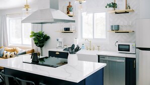 Een moderne keuken met een centraal eiland, witte aanrechtbladen, een roestvrijstalen afzuigkap, een magnetron en een koelkast. Er zijn twee hanglampen, een koffiezetapparaat en een plant op de vensterbank.