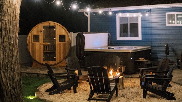 Een hottub met een houten vat, omringd door buitenmeubels en een vuurkuil, verlicht door lichtslingers.