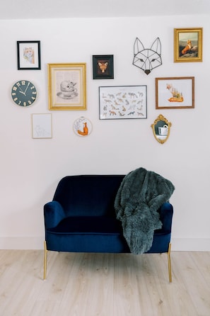 Un sofá de terciopelo azul oscuro con una manta de piel, colocado frente a una pared blanca decorada con varias obras de arte enmarcadas y un reloj de pared.