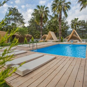 Un área de piscina con muelle de madera, sillas de descanso y una estructura de techo de paja.