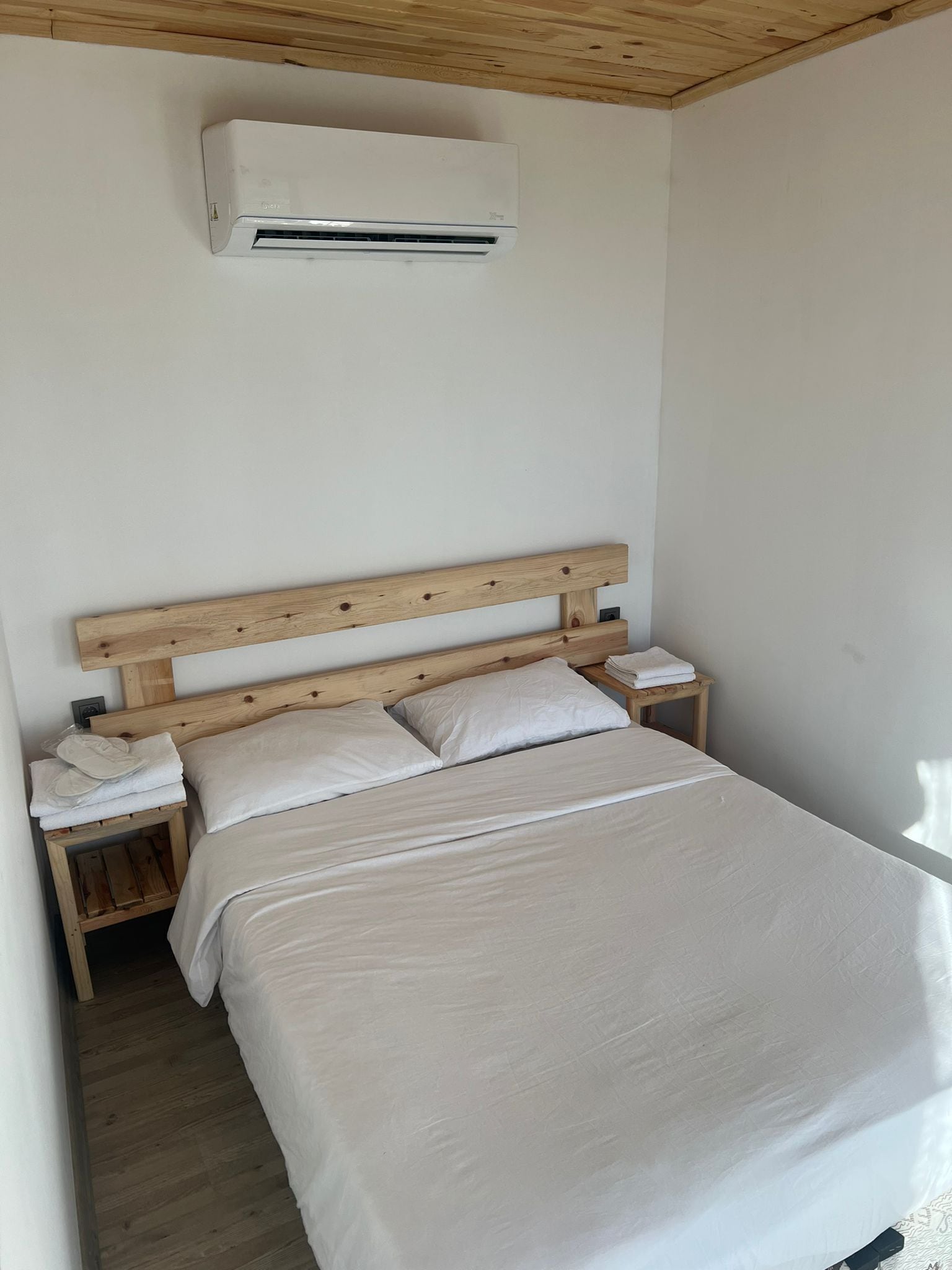 Ein Schlafzimmer mit einem Holz-Kopfteil, einem Einzelbett mit weißer Bettwäsche, einem Nachttisch mit Lampe und einer an der Wand montierten Klimaanlage.