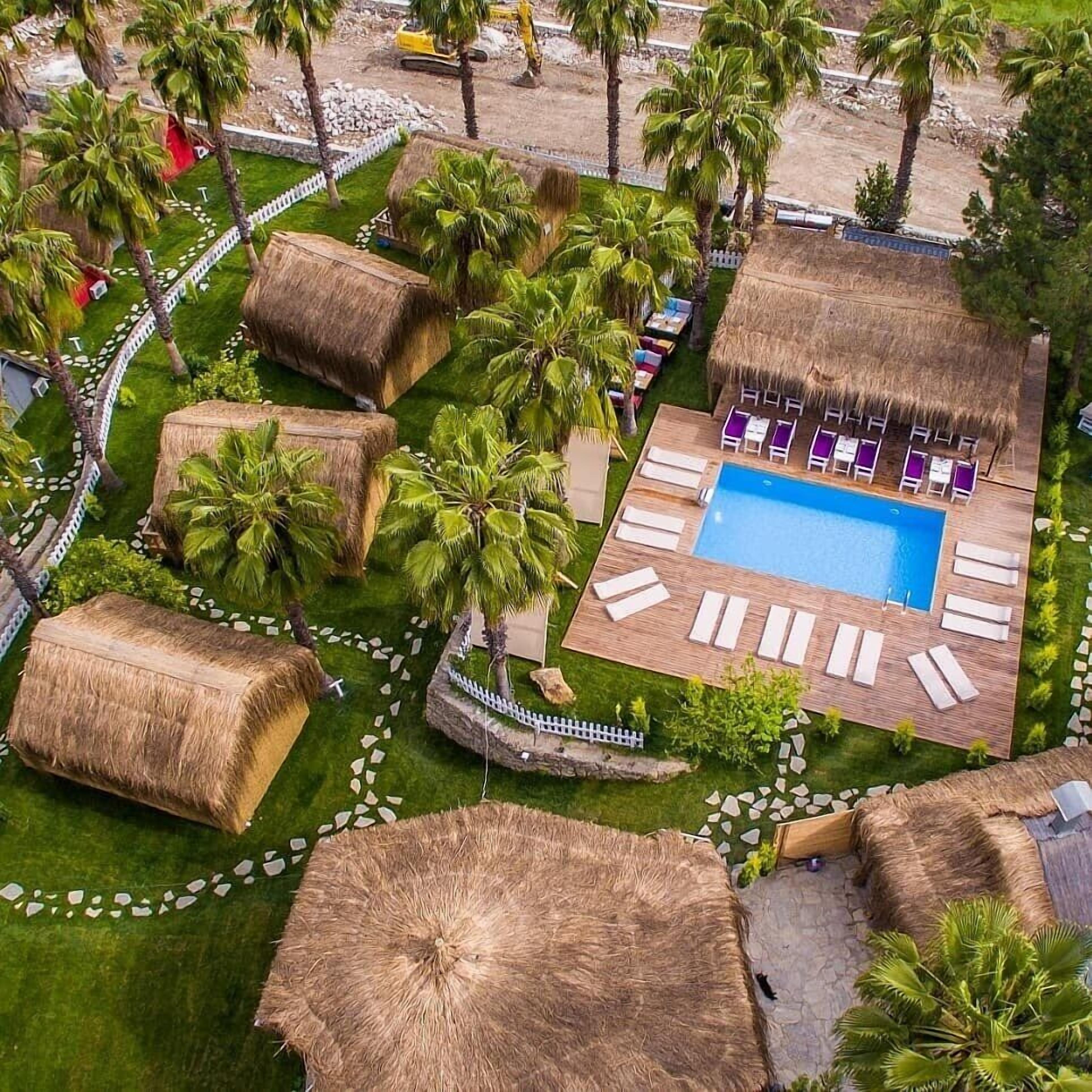 An aerial view of a resort with a swimming pool surrounded by lounge chairs, palm trees, and thatched-roof structures.