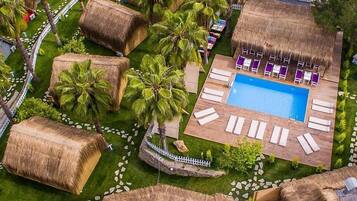 An aerial view of a resort with a swimming pool surrounded by lounge chairs, palm trees, and thatched-roof structures.