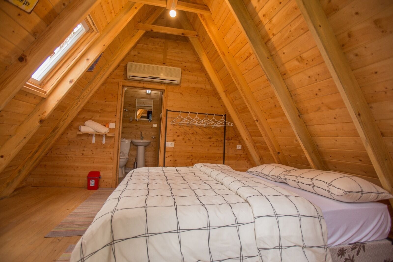 Une chambre mansardée chaleureuse, avec un plafond en pente, un lit recouvert d’une couverture à carreaux, un portant à vêtements suspendu et une petite fenêtre.