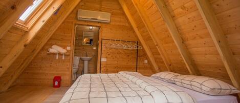 Une chambre mansardée chaleureuse, avec un plafond en pente, un lit recouvert d’une couverture à carreaux, un portant à vêtements suspendu et une petite fenêtre.
