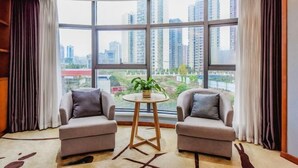 A room with two armchairs, a small round table with a plant, large windows with a city view, and a patterned rug.