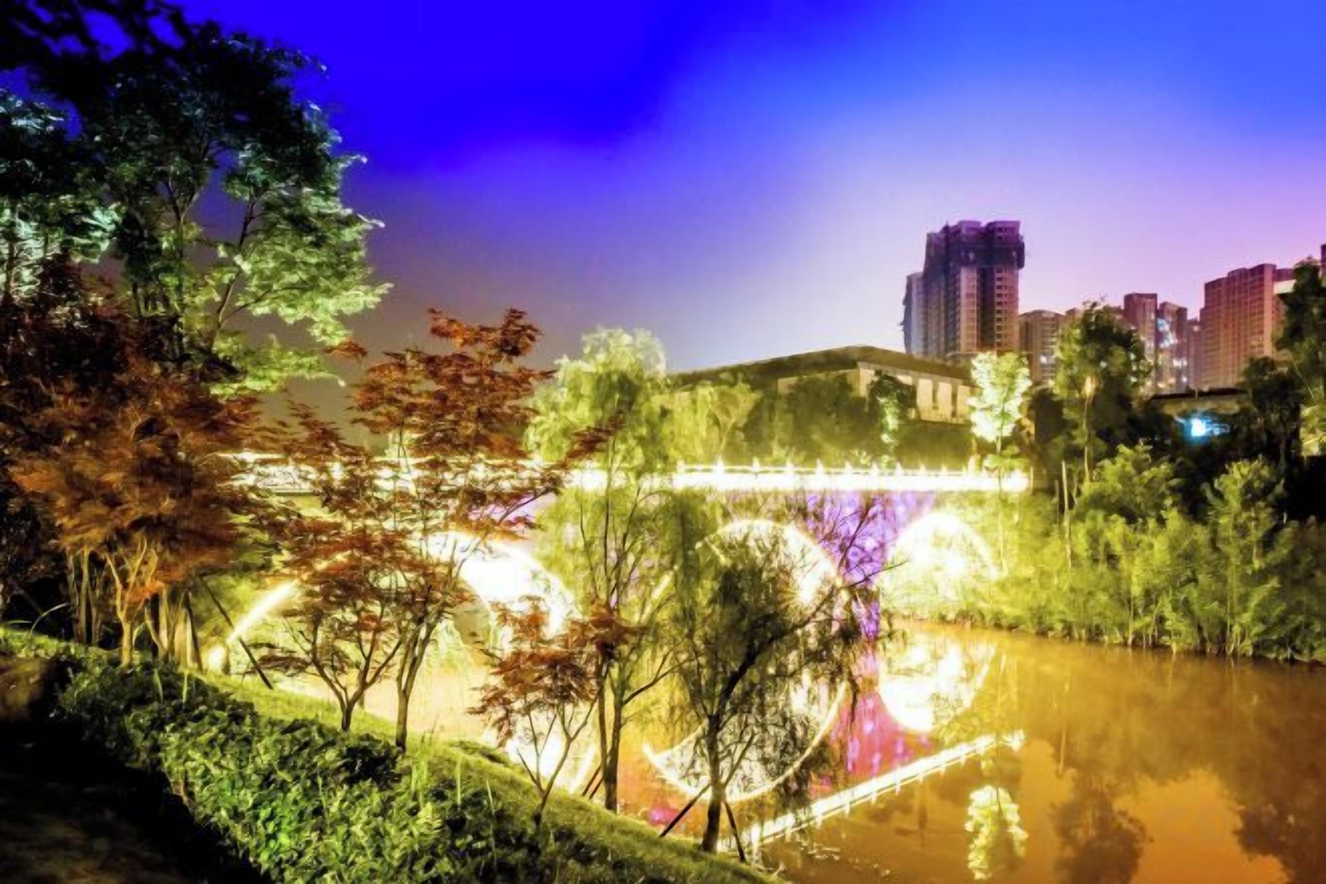 A nighttime view of a riverside park with trees and a bridge.