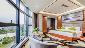 A modern hotel room with a large window, a bed with green pillows, a small round table with a plant, and a view of the cityscape.