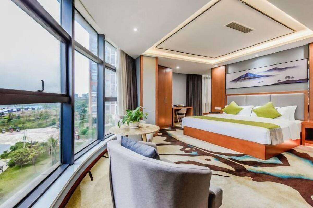 Une chambre d’hôtel moderne avec une grande fenêtre, un lit agrémenté d’oreillers verts, une petite table ronde avec une plante, et une vue sur le paysage urbain.