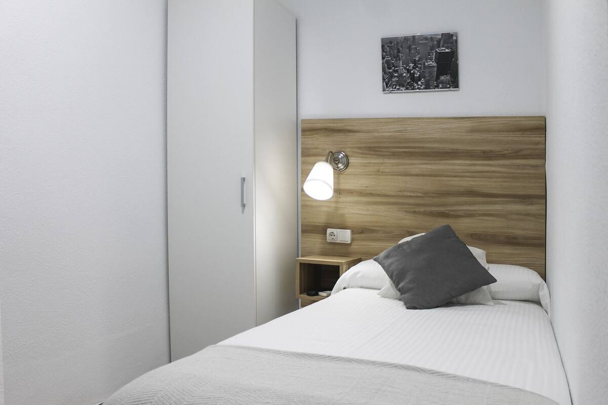 Une chambre d’hôtel moderne avec une tête de lit en bois, une lampe de chevet, une table de chevet et un lit recouvert de draps blancs et agrémenté d’un oreiller gris.