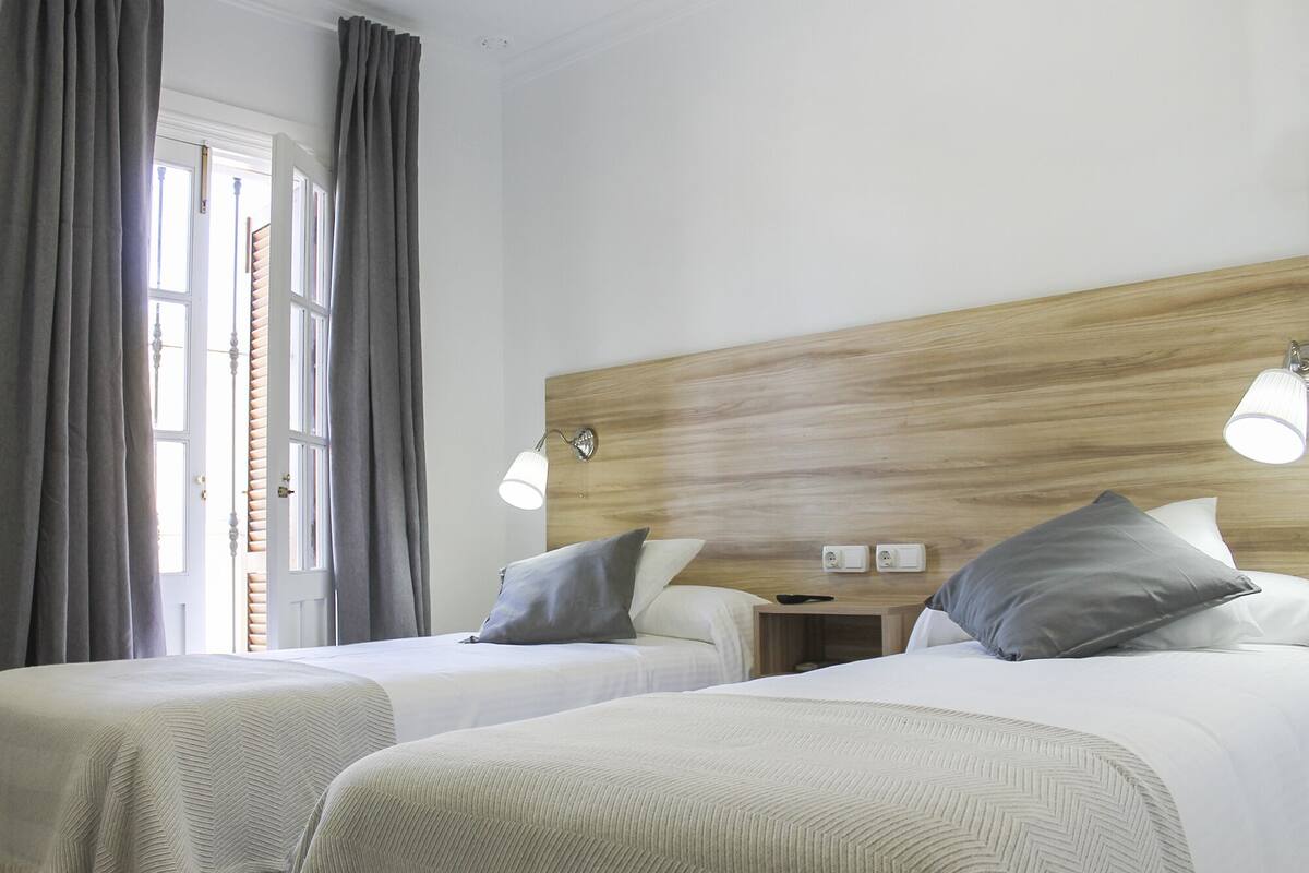 Une chambre d’hôtel avec deux lits, une tête de lit en bois, des lampes de chevet, une table de chevet et une fenêtre avec des rideaux.