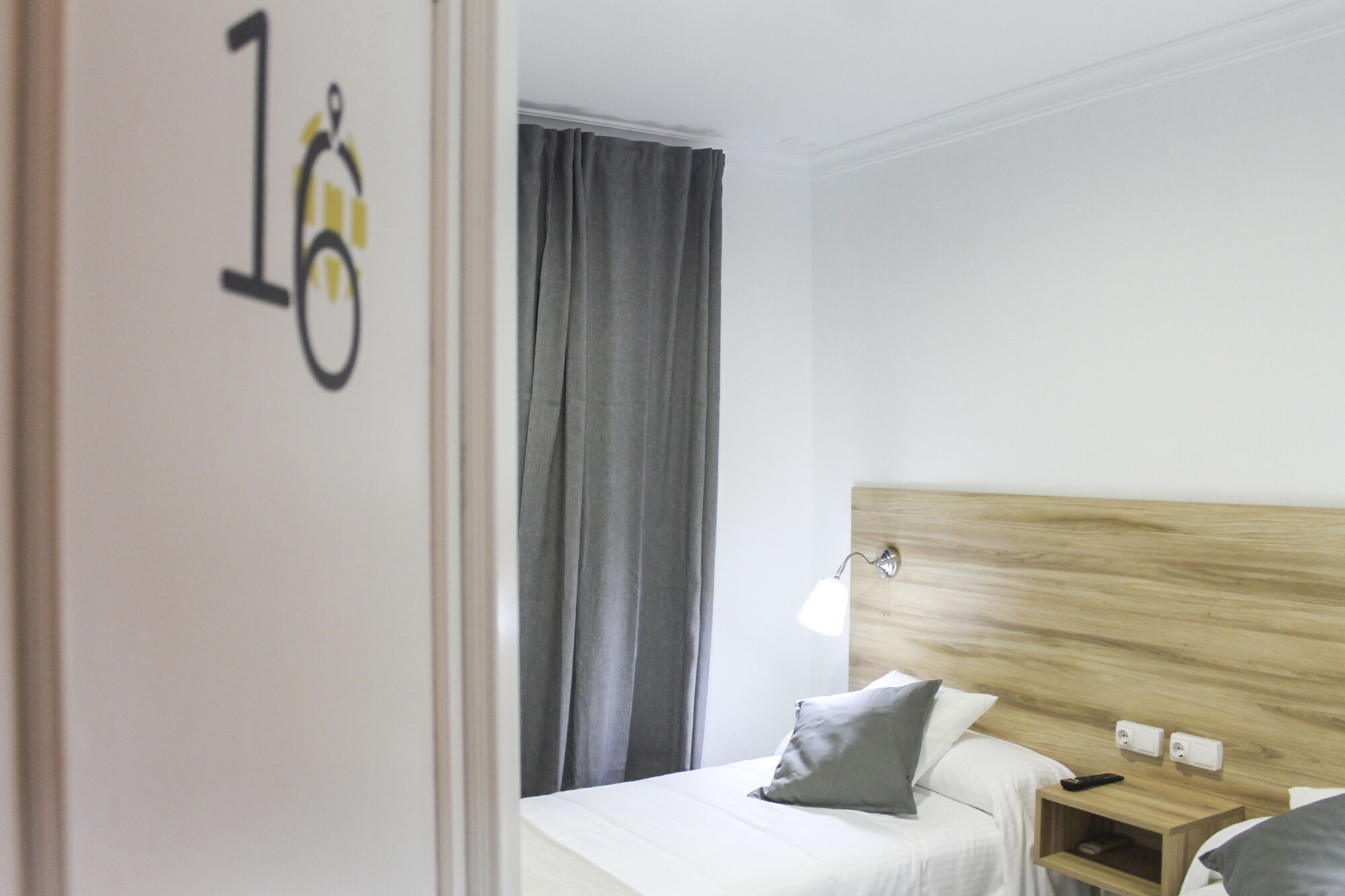 Une chambre d’hôtel avec un lit, une tête de lit en bois, une table de chevet avec une lampe et un rideau gris.