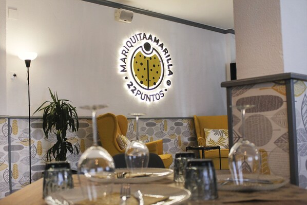 Un comedor con una mesa dispuesta para dos personas, una planta en maceta y un logotipo adherido a la pared de "Marquintamarillamartinos".