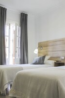 Habitación de hotel con dos camas, cabecero de madera, mesita de noche y ventana con cortinas.