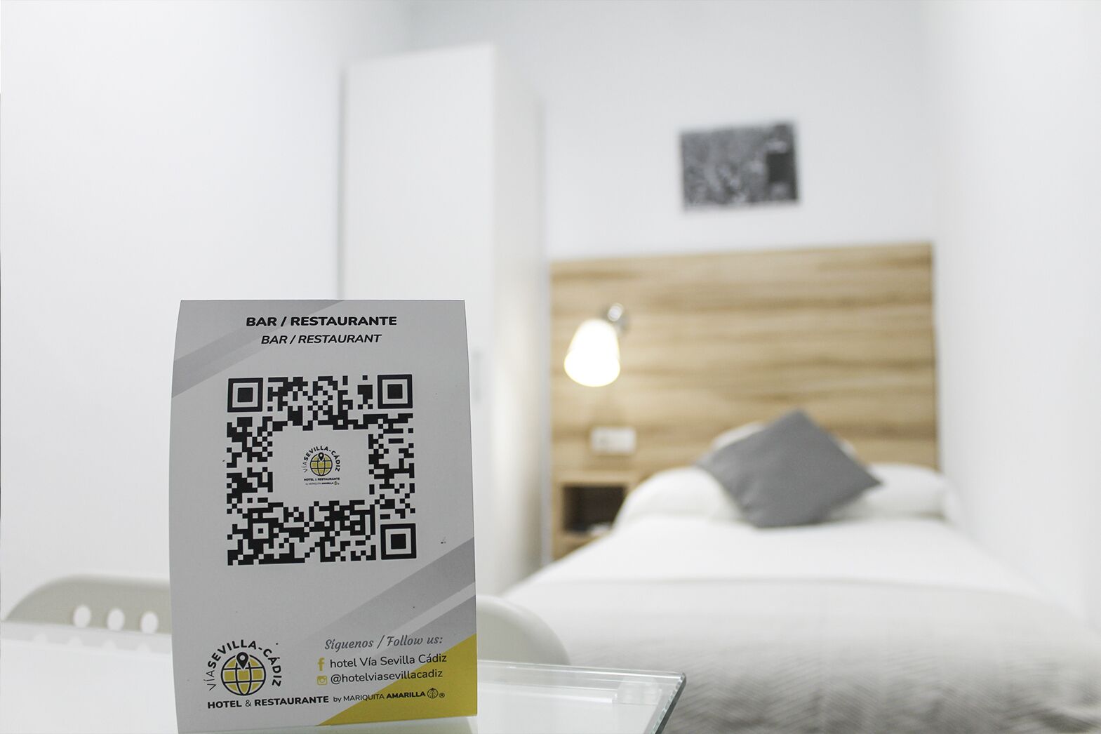 A hotel room with a bed, a bedside table, and a nightstand. There is a QR code on a sign in the foreground.