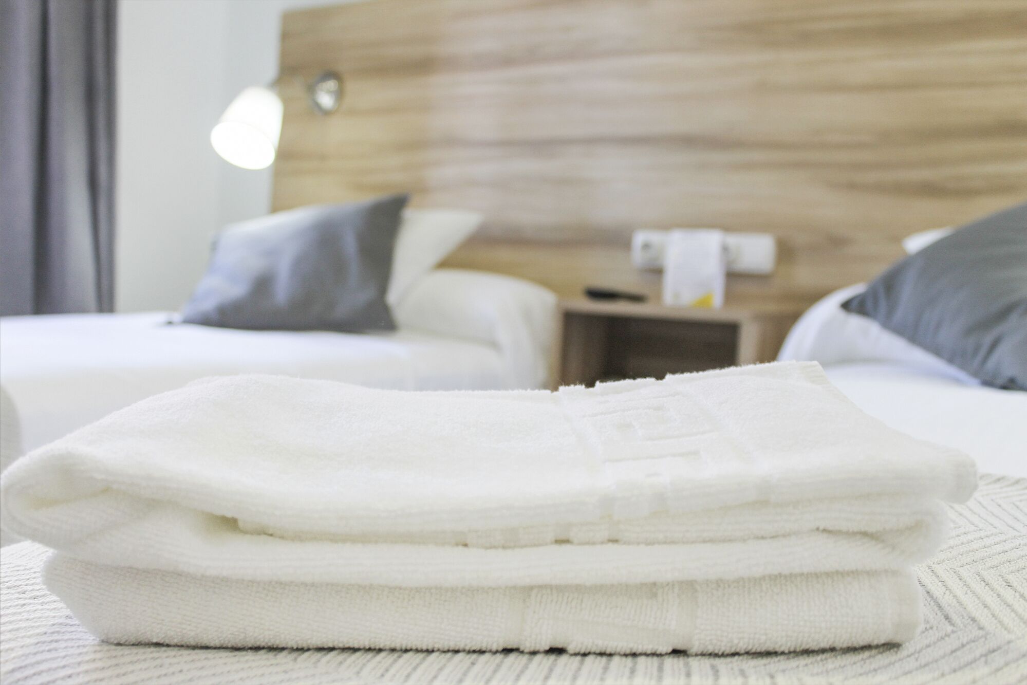 Toalha branca dobrada sobre uma superfície branca texturizada, possivelmente uma cama ou porta-toalhas, em quarto de hotel com cabeceira de madeira e mesa de cabeceira com abajur e controle remoto.