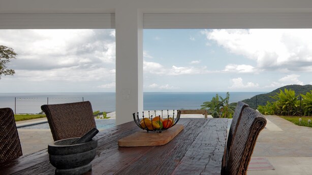 Une table en bois, sur laquelle sont posés un bol en pierre et un panier de fruits, offre une vue imprenable sur la côte, avec ses montagnes et sa végétation luxuriante.