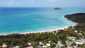 Vue aérienne d’une zone côtière avec une plage, une végétation luxuriante et des bâtiments dispersés.