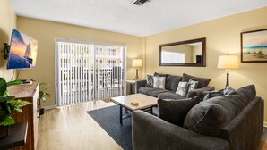 A living room with a sofa set, a coffee table, a TV, and a large window with blinds.