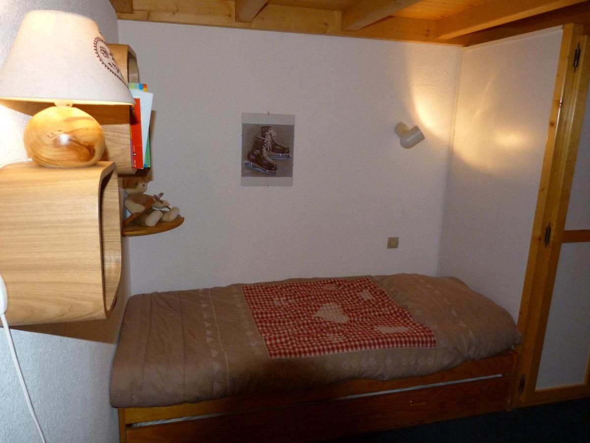 A small, single-bed room with a wooden headboard, a bedside lamp, a wall-mounted shelf with books and a picture, and a red and white checkered blanket.