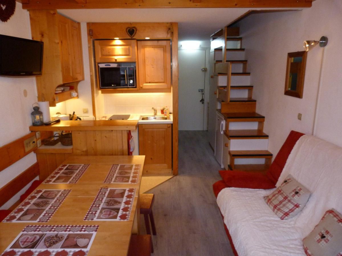 A compact kitchen with wooden cabinets, a microwave, and a sink. A dining table with a placemat is in the foreground. A staircase leads to a bedroom with a white bed and red pillows.