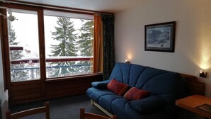 A cozy living room with a blue sofa, wooden chairs, a small table, and a large window with a view of snow-covered trees. - 50m from resort center, 12nd floor, view firs, balcony, tv, 29m², Les Arcs (Les Arcs)