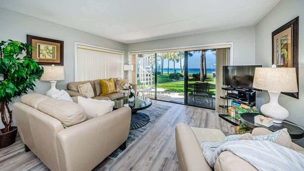 Ein Wohnzimmer mit einer Couch, einem Couchtisch, einem Fernseher und einem großen Fenster mit Blick auf das Meer und Palmen.