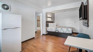 A compact hotel room with a bed, a desk, a chair, a refrigerator, a wall-mounted TV, and a bathroom visible through an open door. - Studio 6 Ontario, Ca - Convention Center - Airport (Ontario)