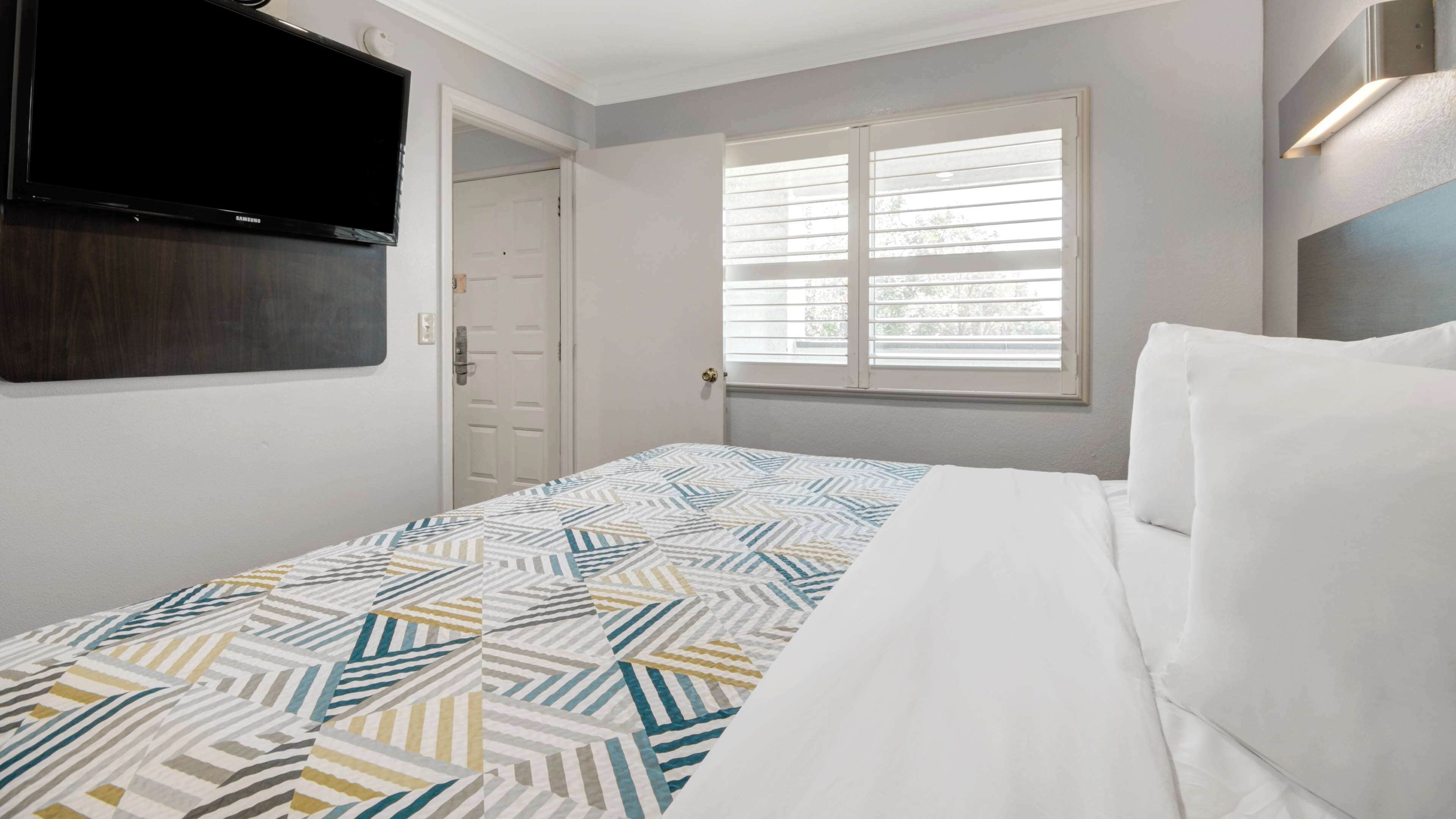 A bedroom with a bed, a TV mounted on the wall, a window with blinds, and a door.