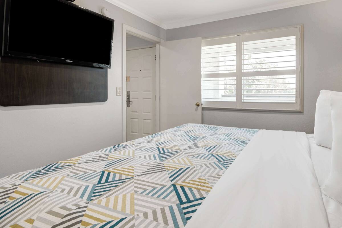 A bedroom with a bed, a TV mounted on the wall, a window with blinds, and a door.