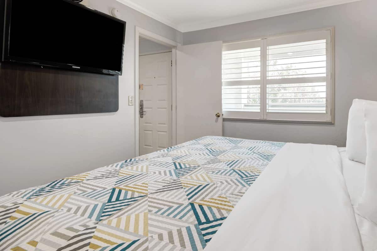Une chambre à coucher avec un lit, un téléviseur fixé au mur, une fenêtre avec des stores et une porte.