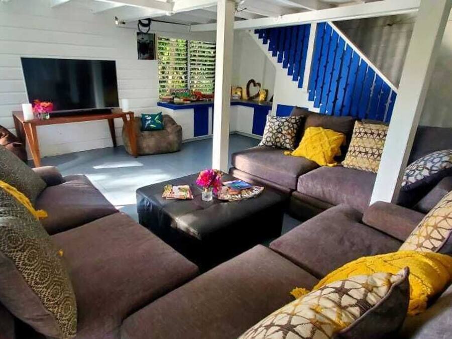 A modern living room with a sectional sofa, a coffee table, and a TV. There is a staircase with blue railings and a white wall with a shelf.