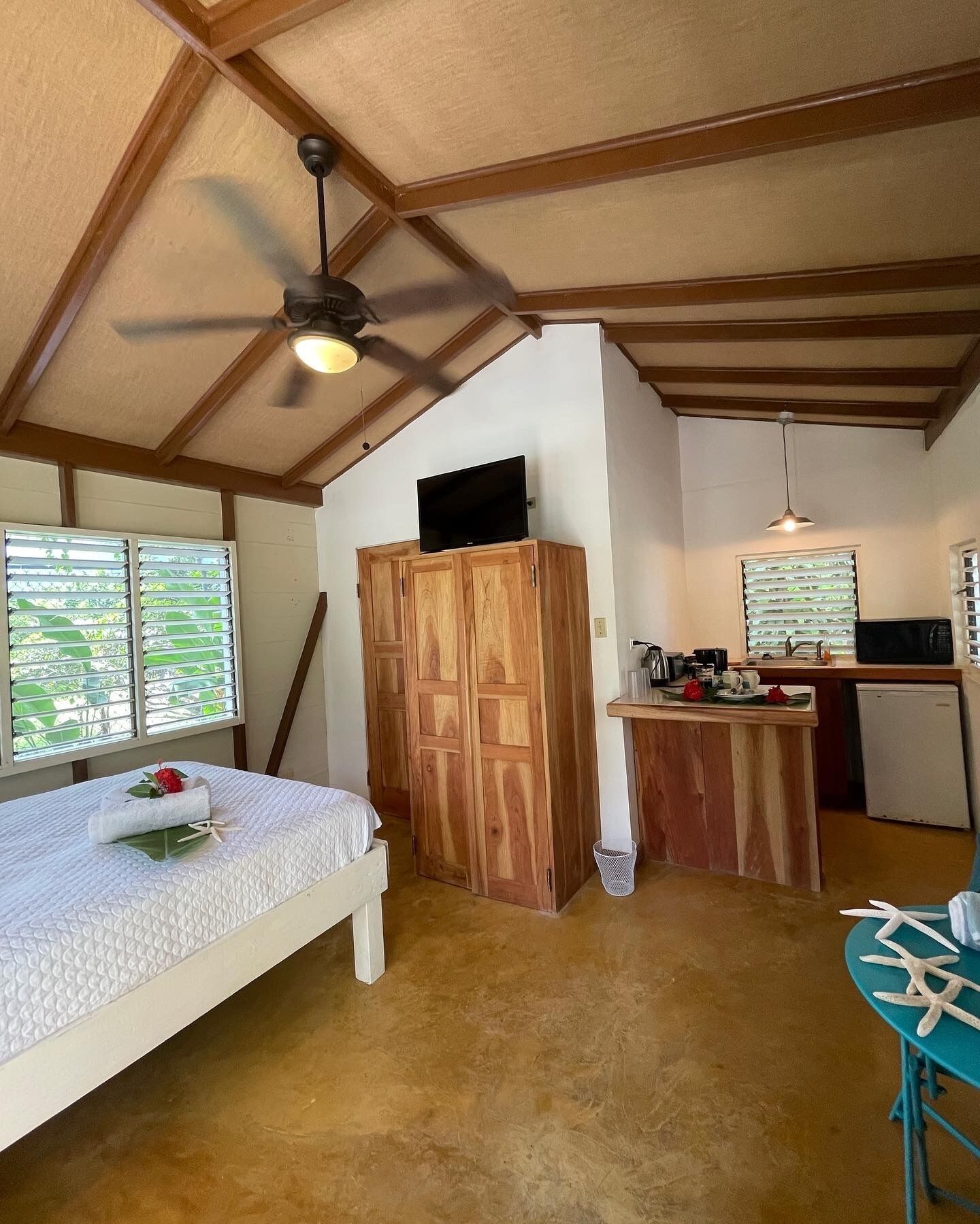 Une chambre avec un lit, une armoire en bois, un ventilateur de plafond, un coin cuisine et une fenêtre avec des stores.