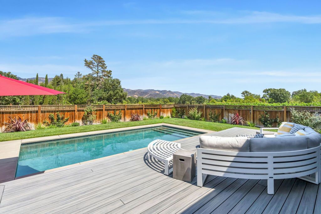 Uma área de piscina com deck de madeira, um sofá branco para área externa e um guarda-sol vermelho.