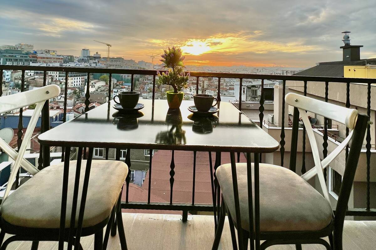 A balcony with a table and chairs, a potted plant, and a view of the sunset over a cityscape.