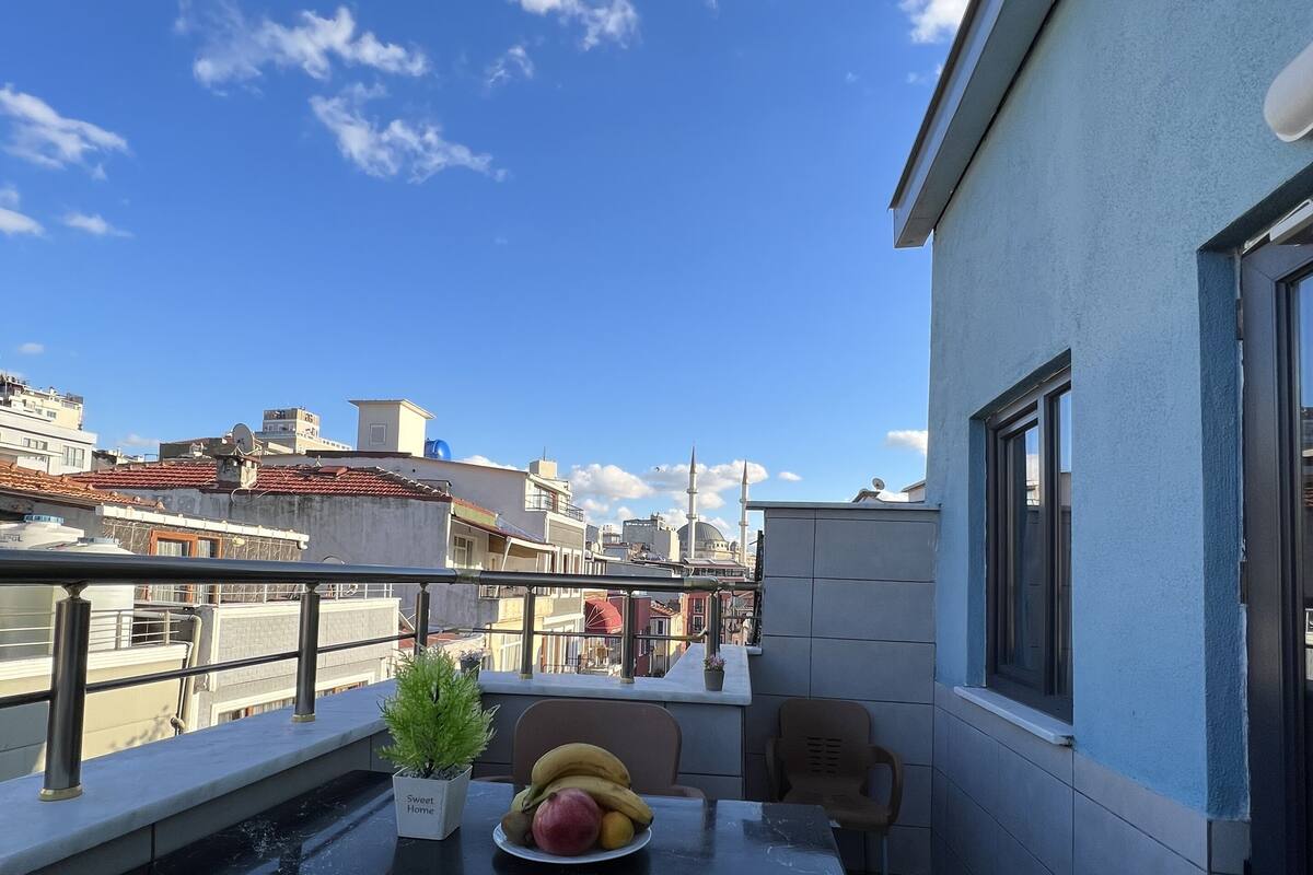 A balcony with a table set for two, a potted plant, and a bowl of fruit.