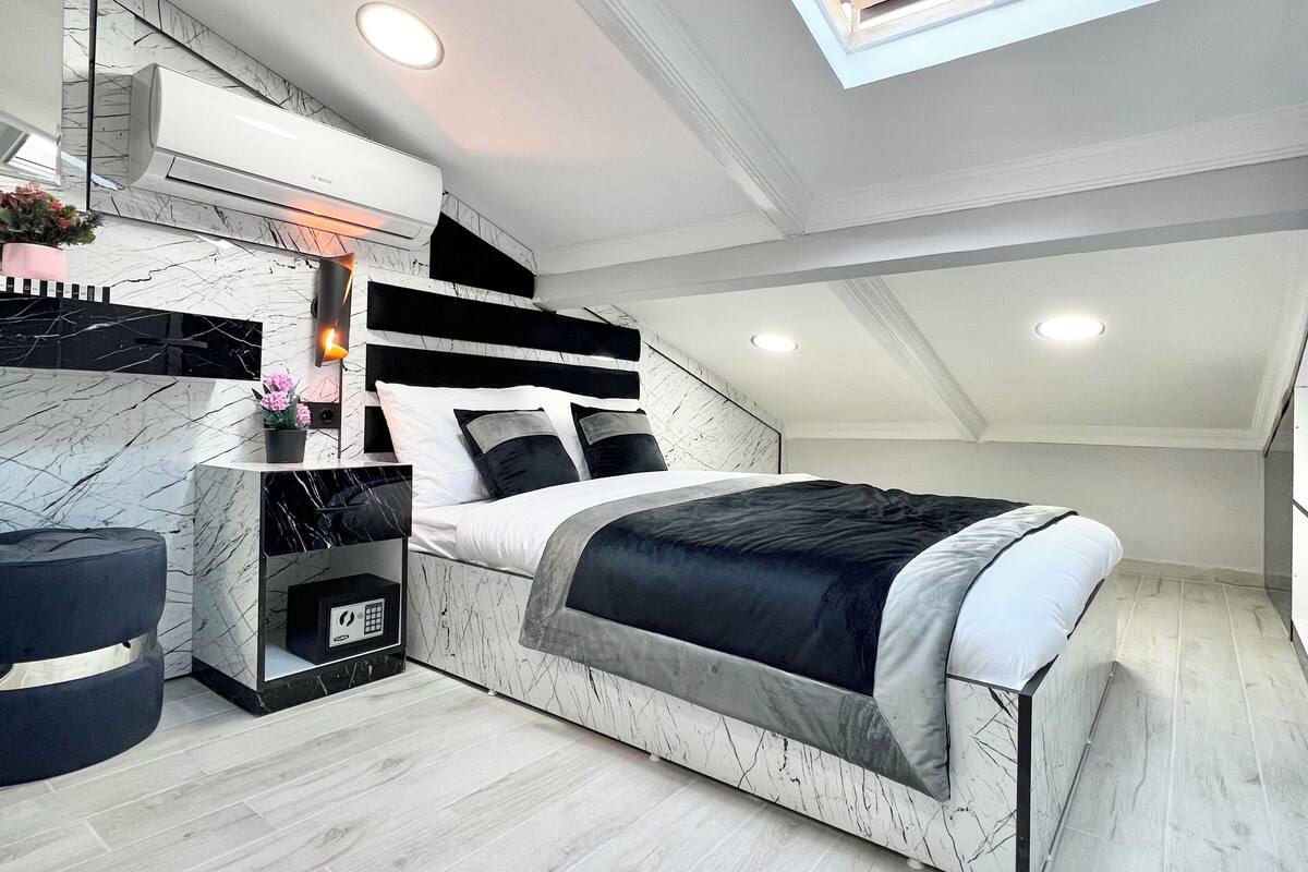 A modern bedroom with a skylight, a bed with a black and white bedspread, a nightstand with a phone, and a marble-patterned wall.