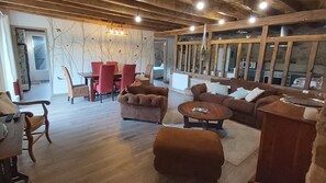 A spacious living room with a wooden ceiling, a dining area with red chairs, and a large window providing natural light.